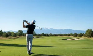 A golfer swings at a tee box with mountains in the background.