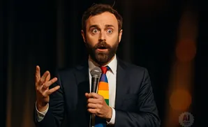 Man in a suit and rainbow tie speaks into a microphone, with his hand raised.
