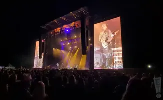 A country singer plays guitar on a large stage with video screens showing his performance to a crowd.