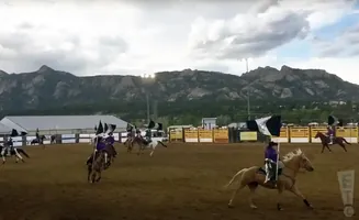 Rodeo riders in purple shirts with flags compete in front of a mountain range.