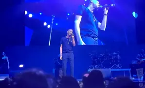 A male singer in a black t-shirt and pants sings into a microphone on a brightly lit stage.