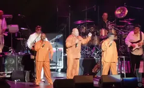 A group of Black men perform on stage in matching orange jumpsuits, singing and playing instruments.