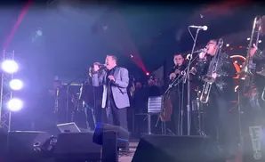 A band performs on stage under bright lights, with a lead singer in a grey suit and a horn section.