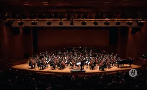 An orchestra performs on a wooden stage in a concert hall, with a pianist at a grand piano.