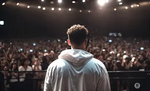 A person in a white hoodie stands facing a large, cheering crowd at a concert, with lights overhead.