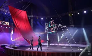 Circus performers in green costumes balance on a metal structure while a large red curtain hangs to the side.
