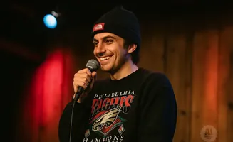 A man wearing a black beanie and sweatshirt with a Philadelphia Eagles logo smiles while holding a microphone.