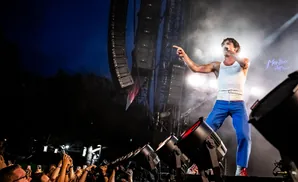Singer in a white tank top and blue pants points to the crowd at a concert, with stage lights and speakers.