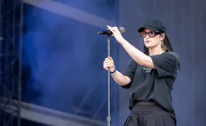 Singer with sunglasses and baseball cap holds microphone on stage.