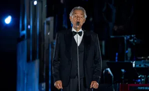 Andrea Bocelli in a tuxedo sings into a microphone on stage.