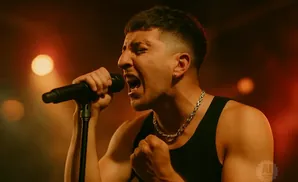 A male singer in a black tank top and silver chain sings passionately into a microphone on a dimly lit stage.