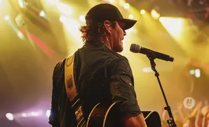 Musician in a black cap and jacket plays guitar on stage, singing into a microphone, with bright yellow and purple stage lights behind him.