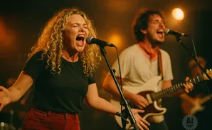 A female singer with curly blonde hair belts into a microphone while a male guitarist plays in the background.