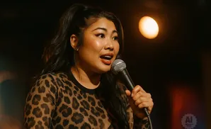 A woman with dark hair pulled into a ponytail wears a leopard-print top and holds a microphone.