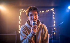 A young man smiles holding a microphone in a dimly lit room with string lights and stage lighting.
