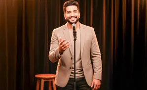 Comedian speaking into a microphone on stage, wearing a beige blazer and dark pants.