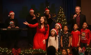 A choir of adults and children in Santa hats sing a Christmas song on stage with a band.