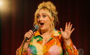 A woman with blonde hair in a messy bun, wearing a colorful shirt, holds a microphone and gestures with her hand while speaking.