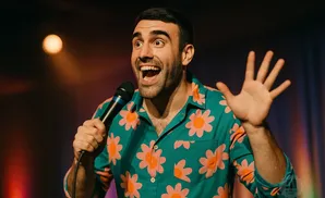 Comedian with a bright floral shirt and microphone raises his hand to the audience.