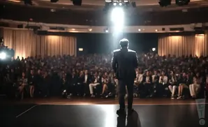 Man in a suit on stage addresses a large, seated audience under bright spotlights.