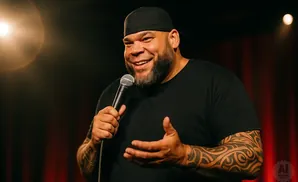 Comedian with beard and tattoos holds a microphone and smiles on stage.