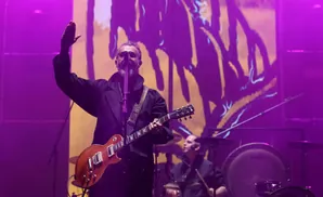 A man with a beard wearing sunglasses and a coat plays a guitar on stage with purple lighting.