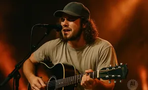 A man in a t-shirt and baseball cap plays an acoustic guitar and sings into a microphone.