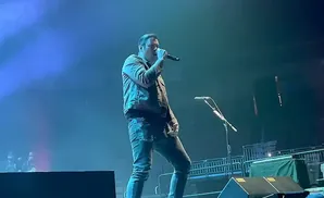 A man sings into a microphone on a dimly lit stage with blue spotlights.