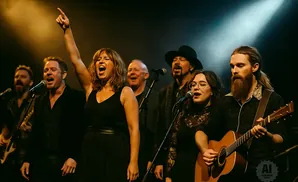 A band performs on stage, with a woman in the foreground singing and pointing upwards.