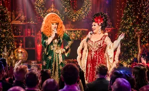 Two drag queens in elaborate gowns perform on a stage with Christmas trees and festive decorations.