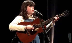 A young person with dark hair sings into a microphone while playing an acoustic guitar on a dark stage.