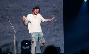A male performer in jeans with red hearts, white t-shirt, smiles and holds a microphone on stage.