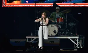 Singer performs on stage in a white outfit with a drummer in the background.