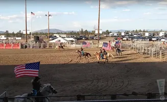 Equestrians ride horses carrying American flags around a rodeo arena, with spectators and vehicles in the background.