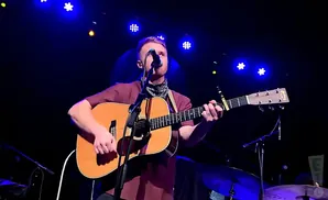 A man in a maroon shirt plays an acoustic guitar on a dimly lit stage with blue lights.
