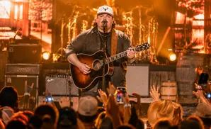 A male country singer plays an acoustic guitar and sings into a microphone on stage, with a crowd in the foreground.
