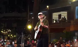 A man in a cap and glasses plays an acoustic guitar on a stage at night.