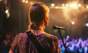Woman on stage with guitar, singing into a microphone with an audience blurred in the background.