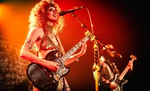 A woman with fiery red hair and dramatic makeup plays a black electric guitar on stage, illuminated by warm, orange stage lights.