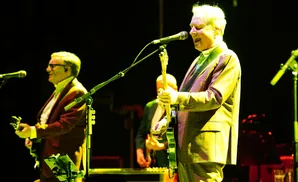 A guitarist in a checkered suit sings into a microphone on stage with other band members behind him.