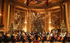 A symphony orchestra performs in a hall decorated with Christmas trees and lights.