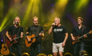 Four men perform on stage: two play guitars, one sings into a microphone, and one plays a bass guitar.