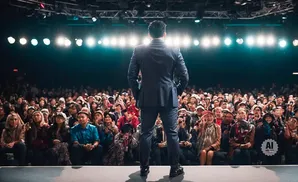 Man in suit on stage facing large audience, lights shine from above.