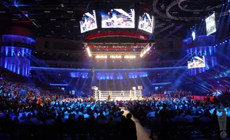 An indoor boxing arena with a brightly lit ring and a large crowd.