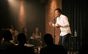 A male comedian tells jokes to a laughing audience in a dimly lit brick room.