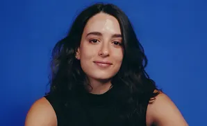 A woman with long, dark hair smiles warmly, looking directly at the camera against a bright blue background.