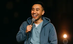 A man in a blue hoodie and scrubs laughs while holding a microphone.