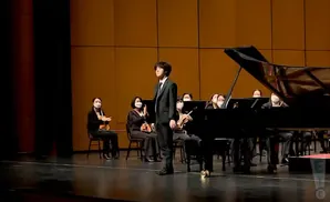 A young man in a suit stands at a grand piano with an orchestra behind him.