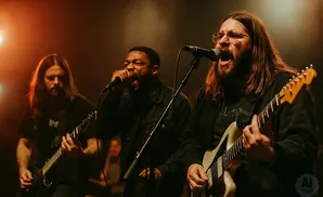 A band performs on stage, with three musicians singing and playing guitars under warm stage lighting.