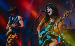 A band performs on stage, with a female bassist and male guitarist in the foreground.